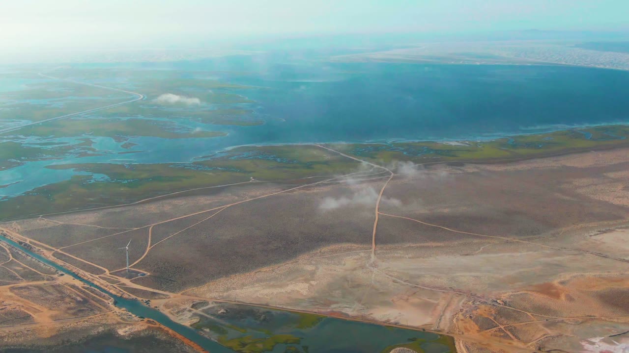 Wide drone view of wetland water, dry terrain, roads, and white mist.