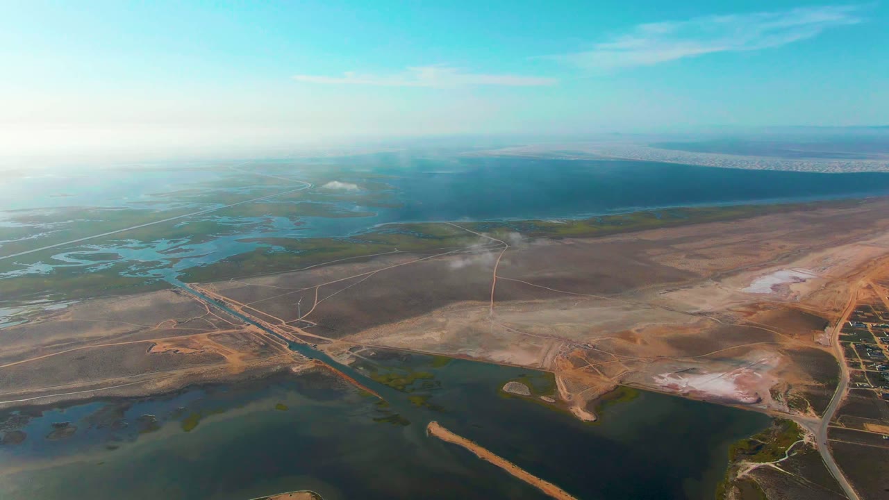 Wide Guerrero Negro aerial frame showing shallow wetland water at left and bottom, brown arid terrain across the center, thin road lines, a faint smoke plume, and no visible people.