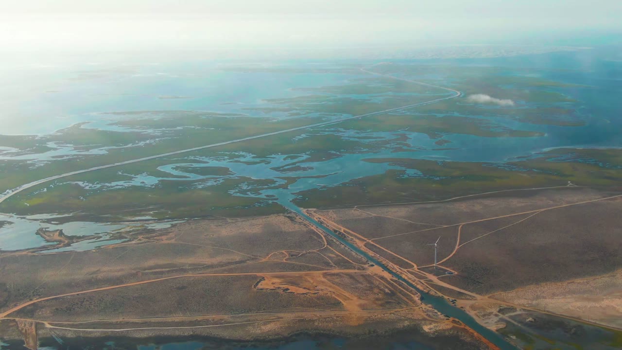 Opening aerial view over Guerrero Negro wetlands with intersecting paved roads, blue-green water channels, brown marsh terrain, distant haze, and no visible people.