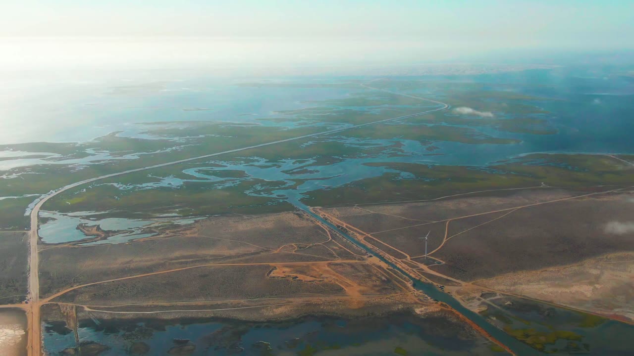 Aerial view in Guerrero Negro with shallow blue water bodies, tan and brown access roads, green marsh patches, bright haze over distant water, and no visible people.