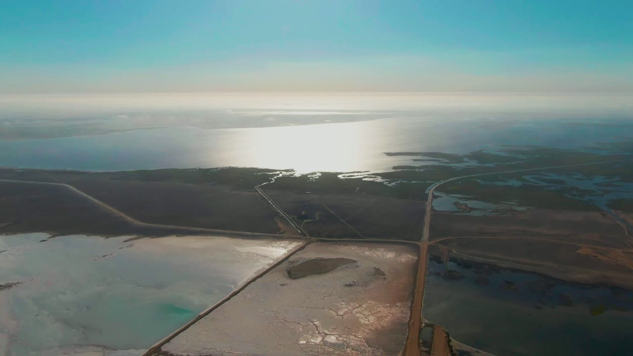 Aerial view over Guerrero Negro salt flats with straight roads crossing dark dry land, shallow water in the foreground, bright reflection on distant water, and no visible people.