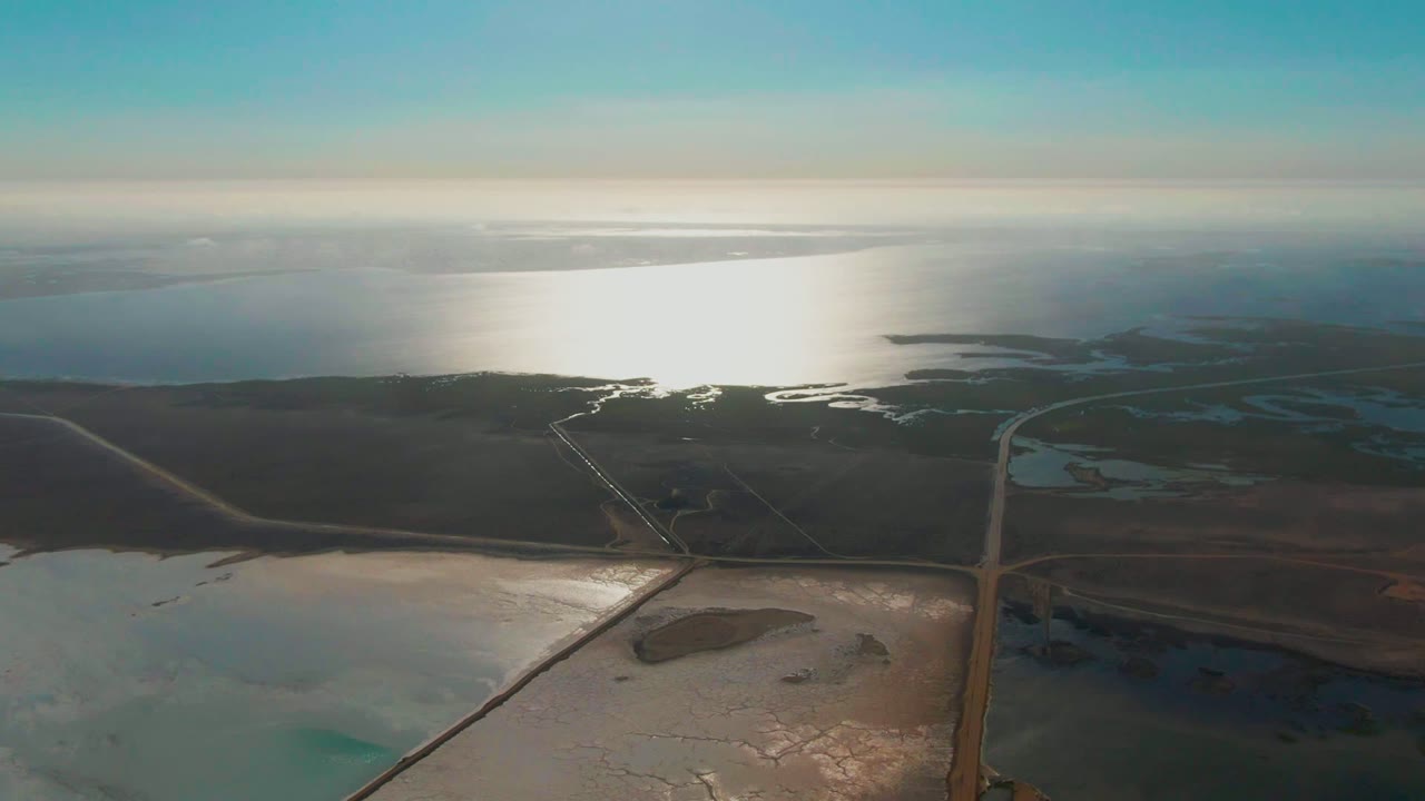 Aerial frame over Guerrero Negro showing divided salt-flat pools below, dark shoreline terrain across the middle, bright reflected sunlight on open water, and no visible people.