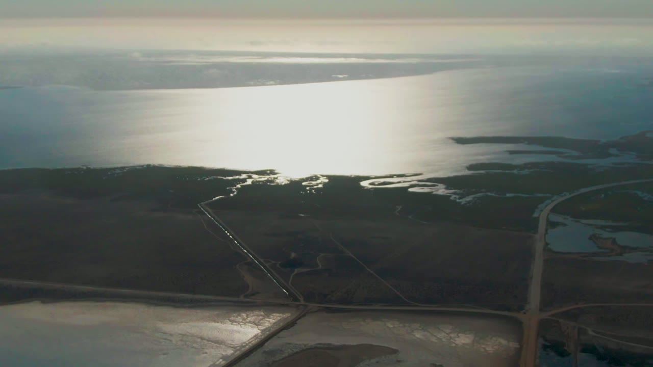 Aerial frame over Guerrero Negro wetlands with a straight road crossing dark flats, reflective ocean in the background, shallow water channels, and no visible people.
