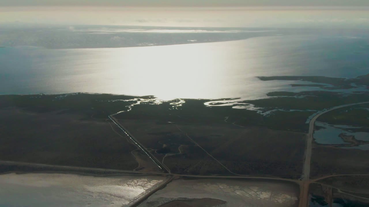 Drone view across dark wetland flats toward reflective coastal water.