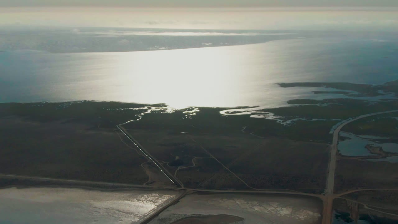 Wide aerial view of Guerrero Negro coastal marsh channels beside a straight road, with shimmering water and a hazy shoreline in the distance and no visible people.