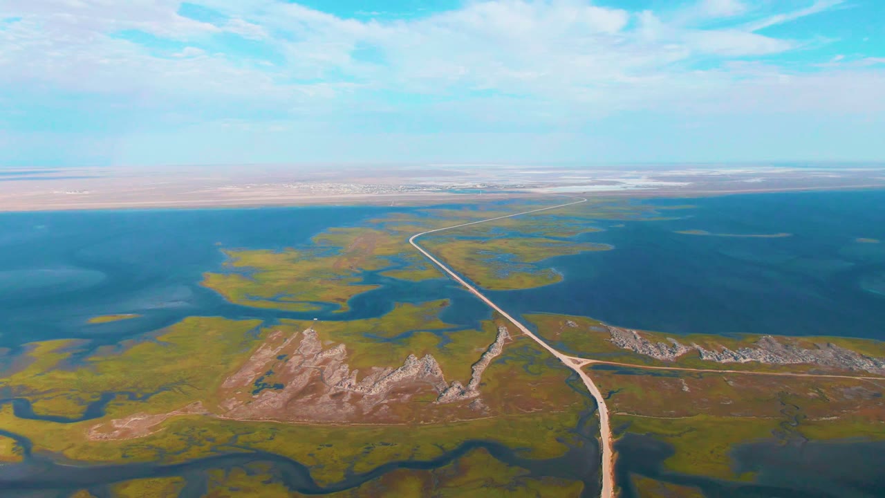 Dirt road bends through green wetlands toward a large blue water body.