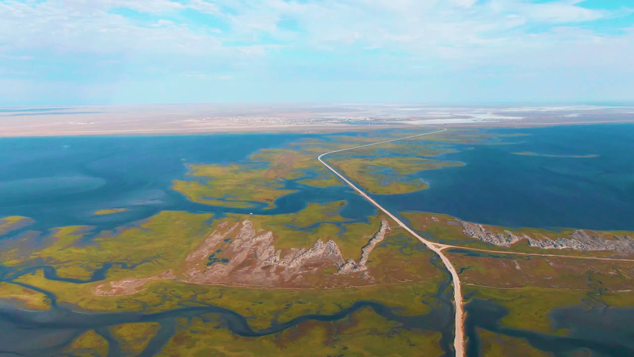 Aerial view near Guerrero Negro, Baja California Sur, with a pale road entering from the lower center, green marsh and blue water on both sides, low rocky ridges midframe, and no visible people.