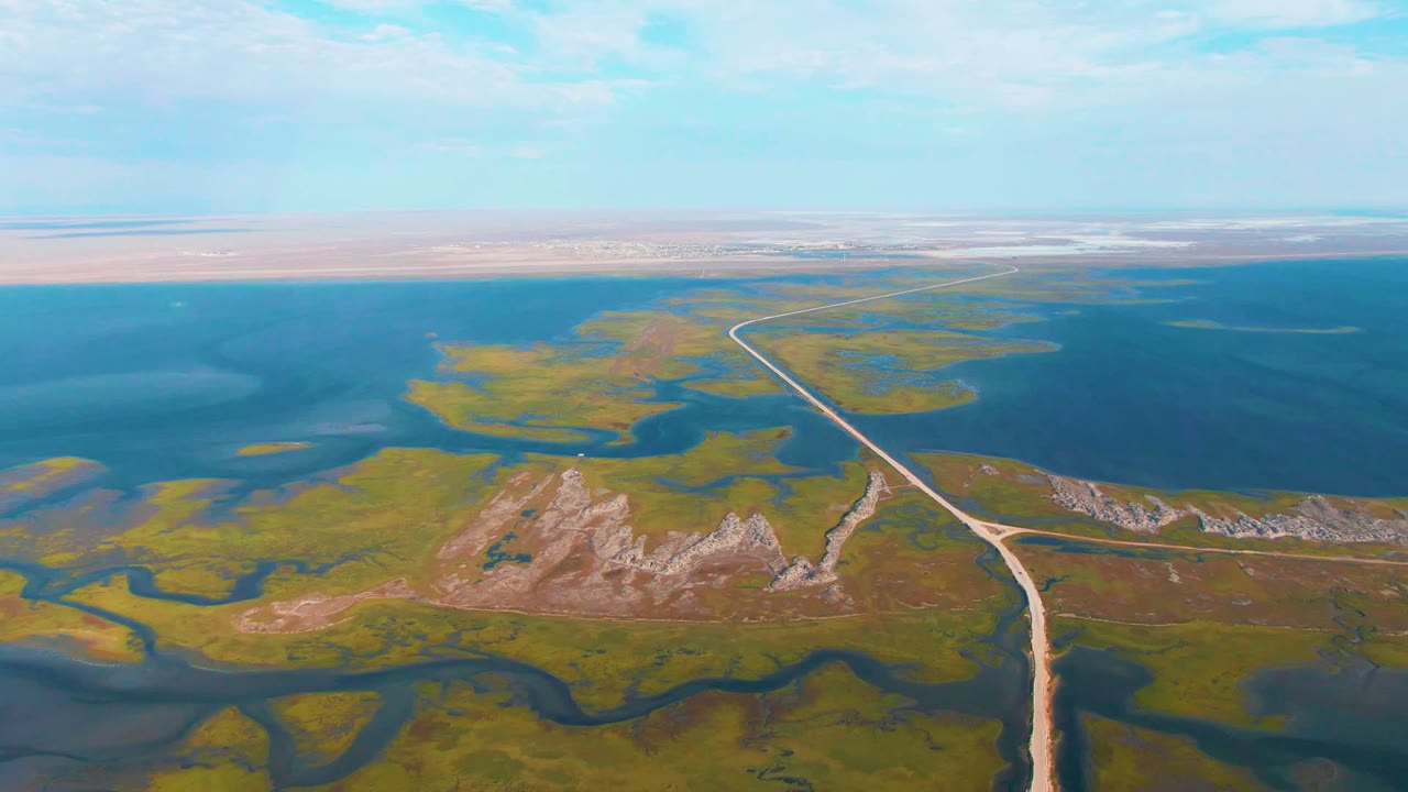 Wide aerial frame near Guerrero Negro, Baja California Sur, with wetland channels in the foreground, a pale road leading toward the horizon, blue water across the upper frame, and no visible people.