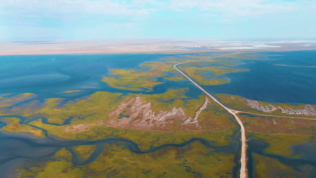 Opening aerial frame over Guerrero Negro wetlands, showing a pale dirt road running through green marsh, blue water channels, rocky low hills, distant desert horizon, and no visible people.