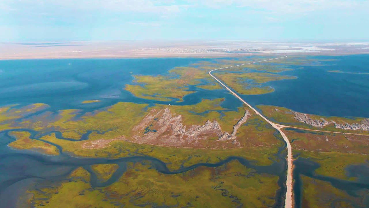 Wide frame over Guerrero Negro wetlands, with a winding dirt road leading toward the horizon, irregular green marsh islands, blue water, low hills, and no visible people or vehicles.