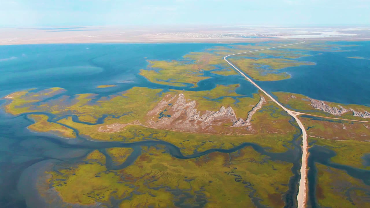 Aerial frame from Guerrero Negro, Baja California Sur showing a pale road cutting between green marsh, blue lagoon water, rocky tan ground, and no visible people.