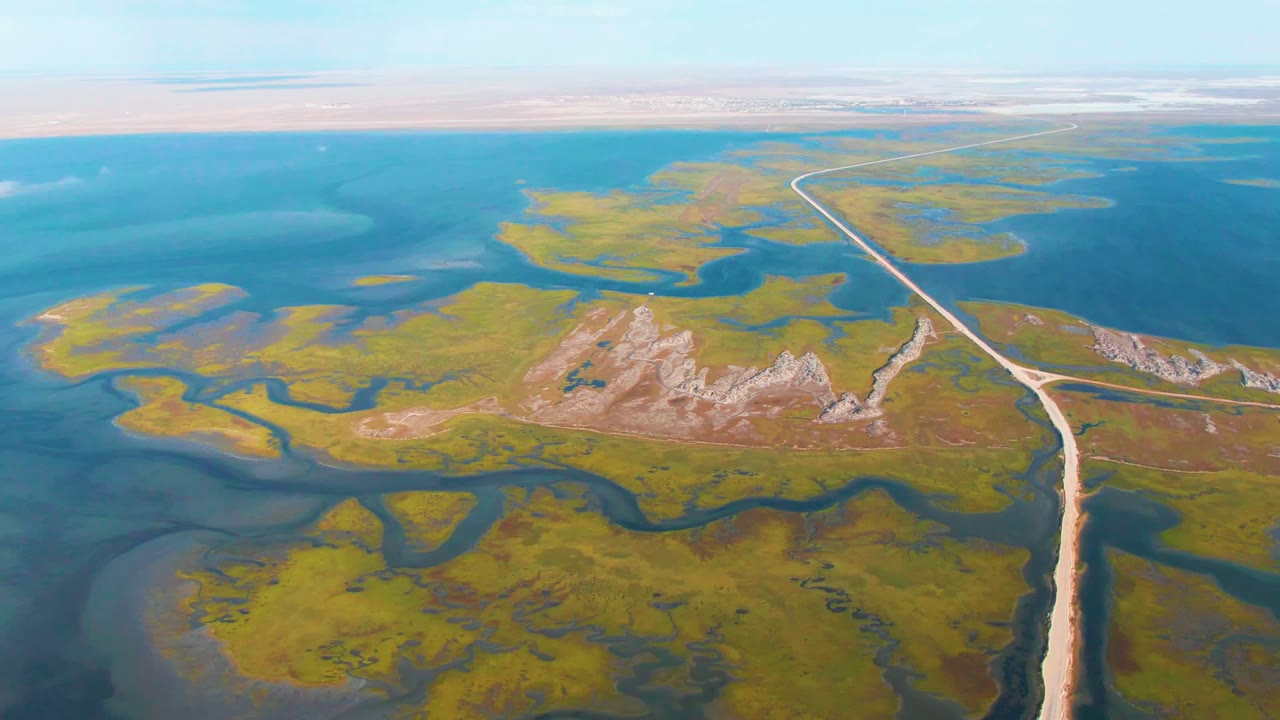 Wide aerial frame from Guerrero Negro showing the long road bending across marshland toward distant water, with tan rocky patches and no visible people.