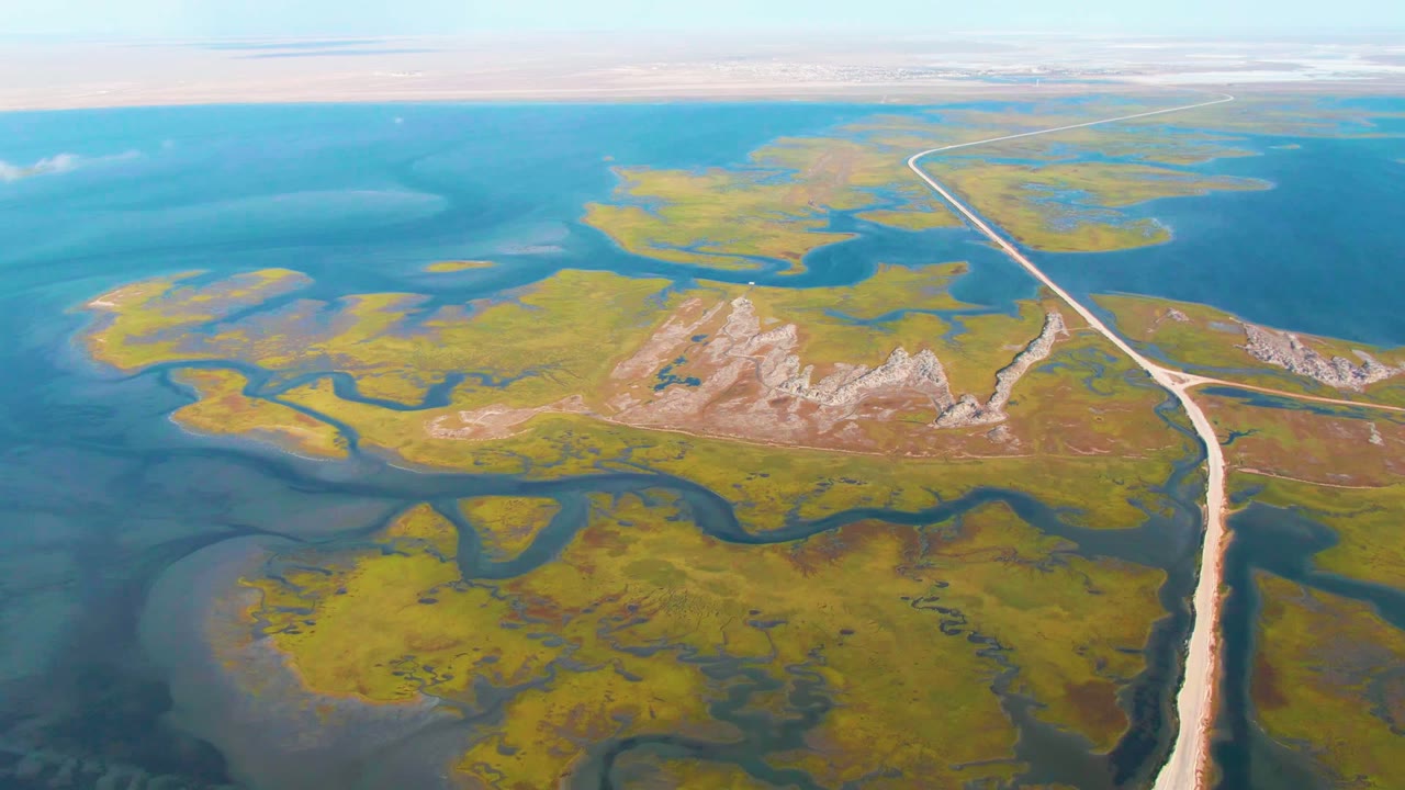 Aerial view over Guerrero Negro wetlands showing blue water channels around green marsh patches, pale rocky ground near center, a dirt road at right, and no visible people.