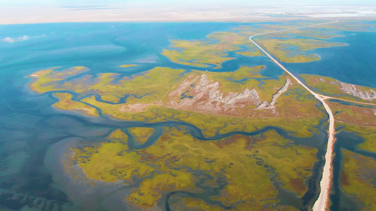 Aerial frame over Guerrero Negro wetland terrain with a long pale road along the right edge, blue water beyond, green marsh shapes below, exposed pale land near center, and no visible people.