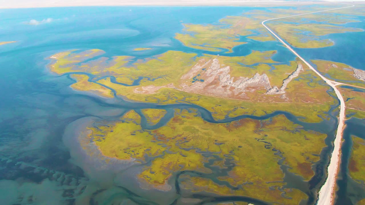 Opening aerial frame over Guerrero Negro, Baja California Sur, with yellow-green marshland, turquoise water, dark winding channels, a pale road at right, and no visible people.