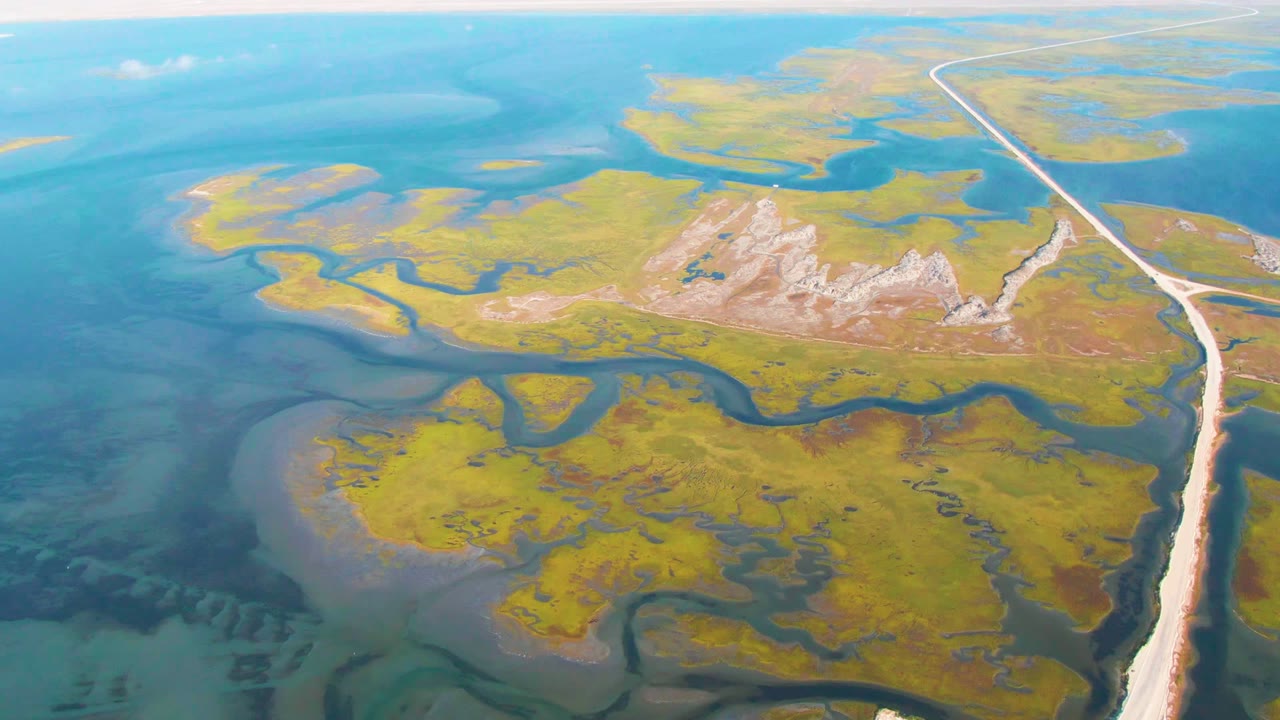 Coastal wetland from above with marsh patches, shallow blue water, winding channels, and road.