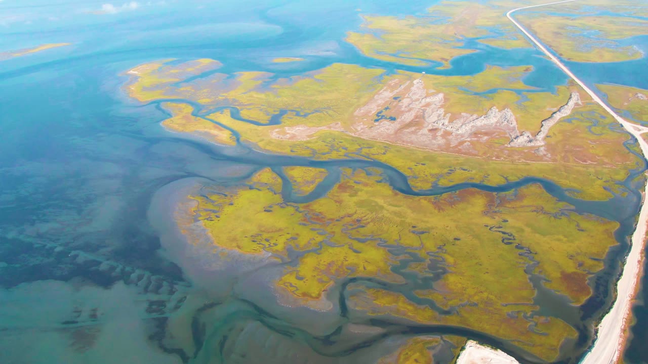 High aerial view of marsh islands and blue water with a road running along the right side.
