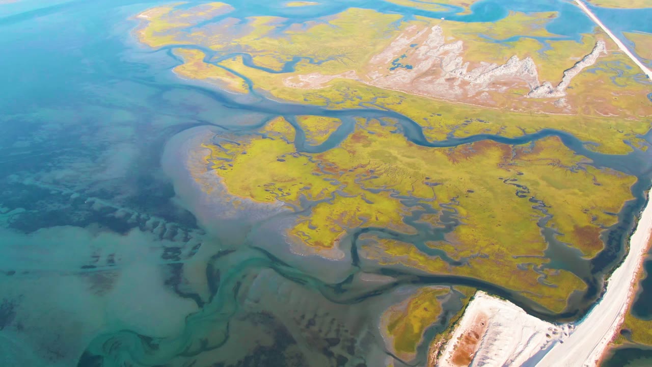 High aerial frame over Guerrero Negro, Baja California Sur, with turquoise water, yellow-green land patches, pale shoreline edges, and no visible people.