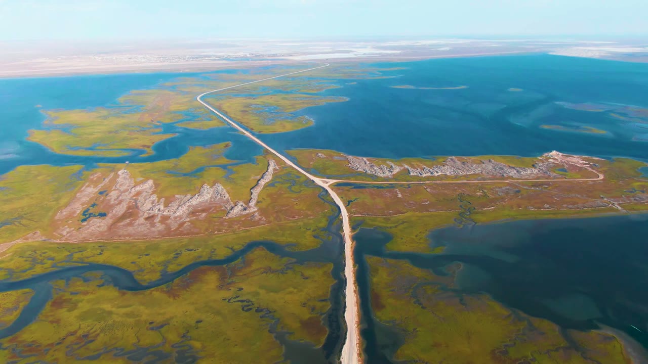 Aerial frame over Guerrero Negro wetlands showing a pale road leading through green and brown vegetation toward blue lagoon water, low pale hills, a hazy horizon, and no visible people.