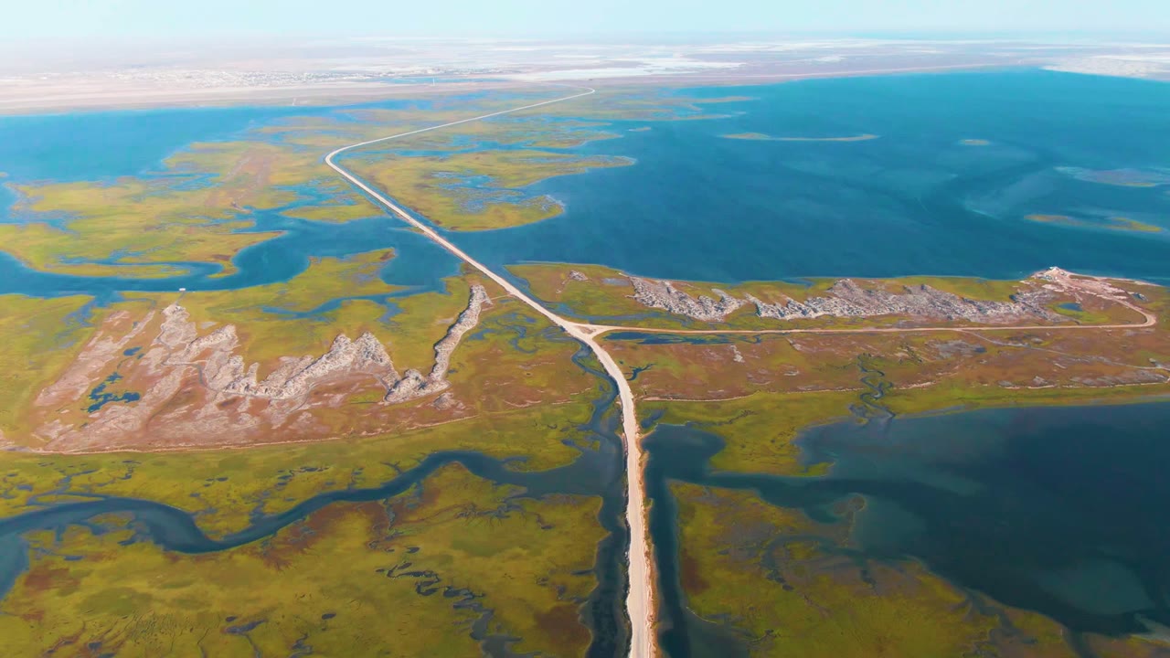 High aerial frame over Guerrero Negro showing the road angled from the lower edge toward distant wetlands, a pale rocky ridge to one side, expansive blue water, and no visible people.