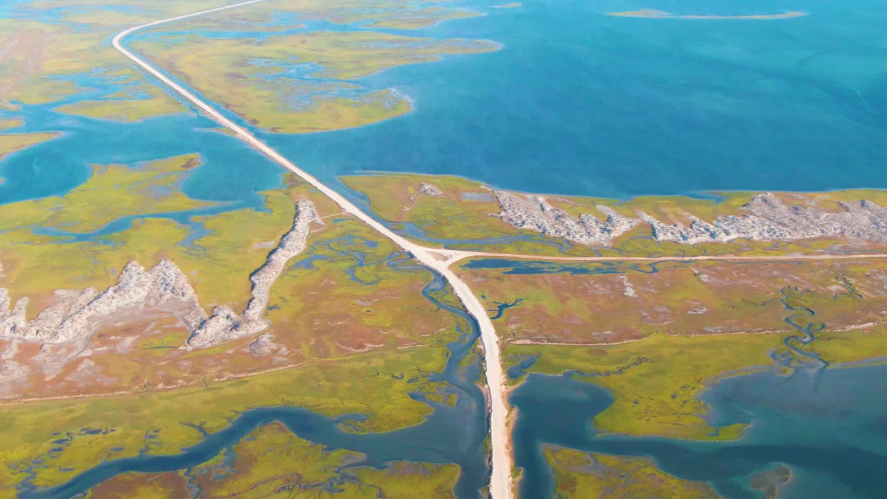 Aerial view in Guerrero Negro, Baja California Sur, with a pale road running diagonally across blue water, green wetlands, low rocky hills, and no visible people.