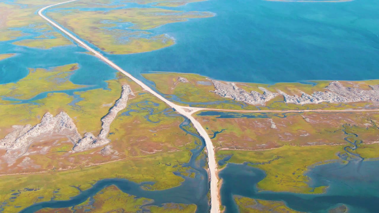 Drone view of a straight wetland road beside blue water and rocky hills.