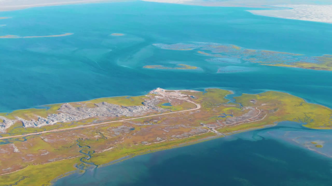 Aerial frame over Guerrero Negro, Baja California Sur, showing a low island with a pale runway, green-brown marshland, blue coastal water, distant pale white landforms, and no visible people.