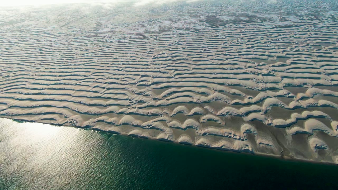 High aerial view of rippled sand flats beside dark green water in Guerrero Negro, Baja California Sur, Mexico, with bright water reflection and no visible people.