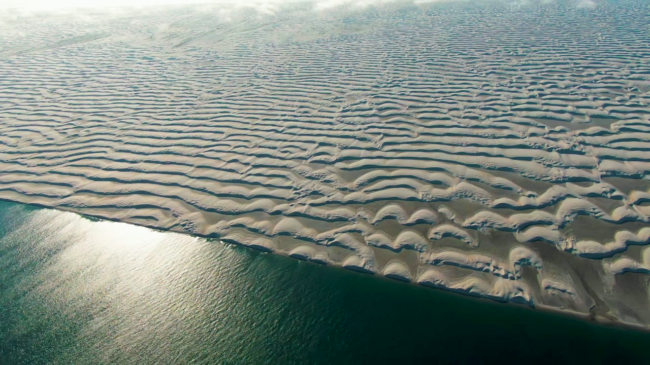 Opening aerial view in Guerrero Negro, Baja California Sur, Mexico, with rippled pale sand dunes above dark green shoreline water and no visible people.