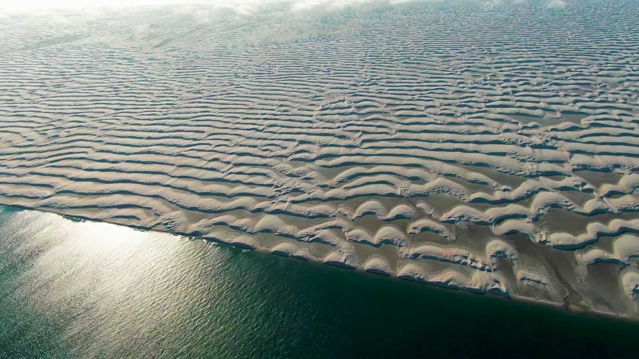 High angled view in Guerrero Negro with pale rippled dunes covering the upper frame and calm green ocean along the lower edge, with no visible people.