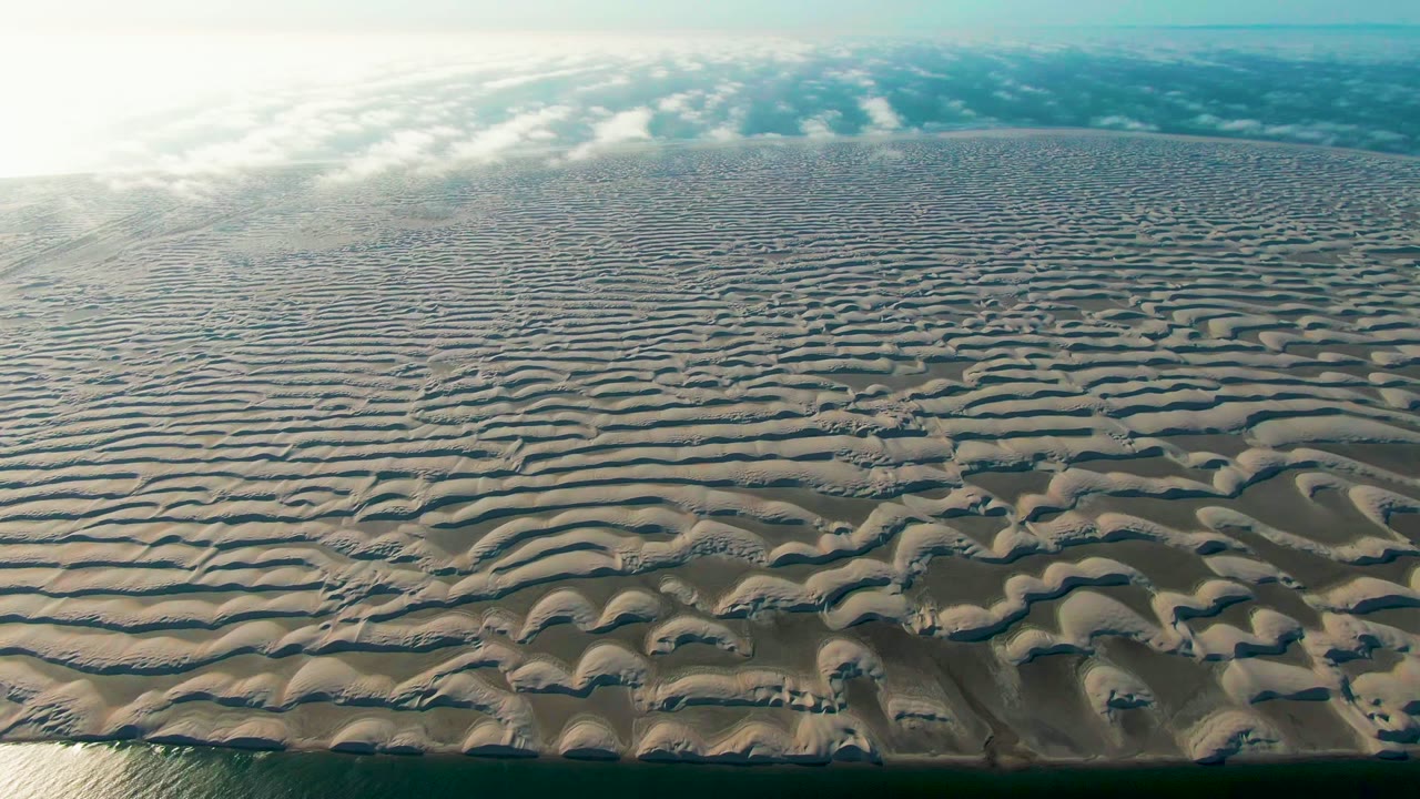 Opening aerial frame in Guerrero Negro, Baja California Sur, showing rippled wet sand stretching toward a hazy blue-white sky, with shallow water at the lower edge and no visible people.