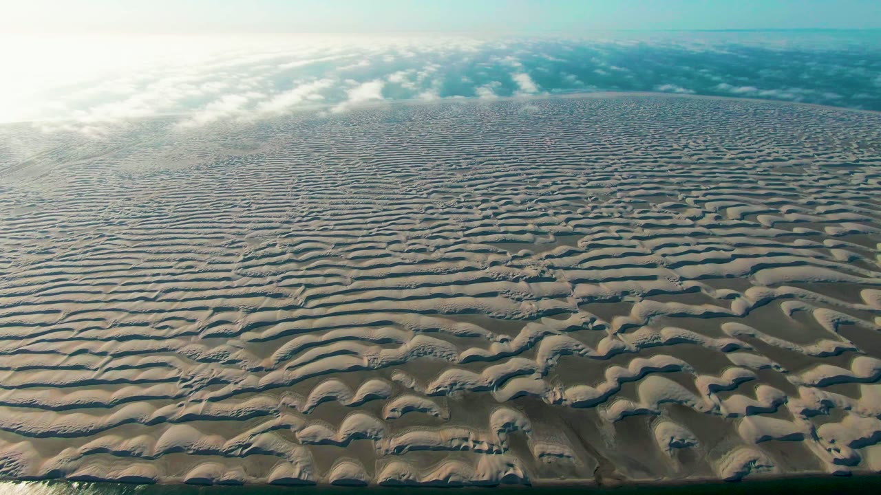 At 0:02, the aerial view in Guerrero Negro holds on repeating sand ridges, darker wet channels, a bright hazy sky band, and no visible people or structures.