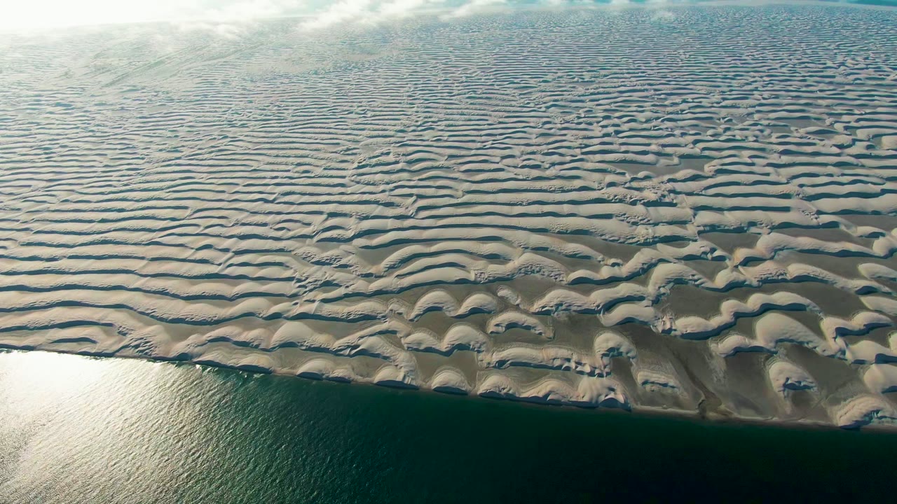 Aerial frame over Guerrero Negro, Baja California Sur, with wavy sand dunes spread across the view, ocean water along the lower edge, pale clouds near the top, and no visible people.