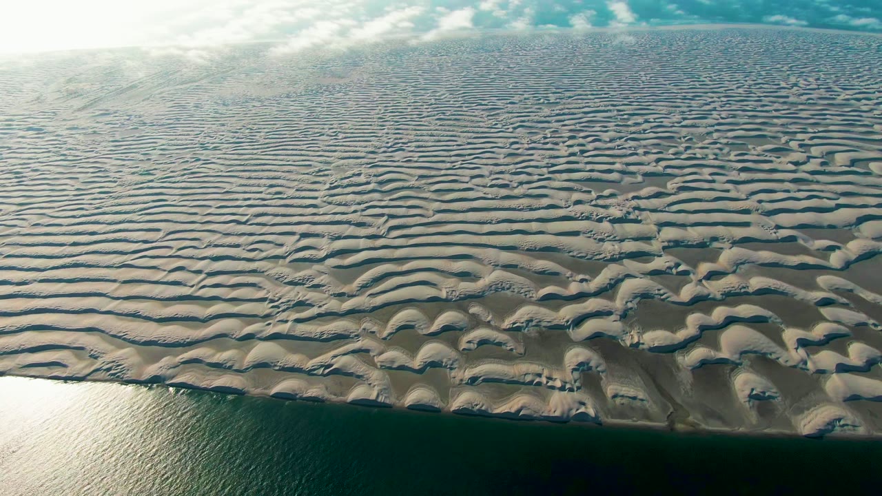 The Guerrero Negro aerial view holds on repeating sand waves, rounded dune mounds near the lower center, a narrow ocean band at the bottom, bright cloud cover above, and no visible people.