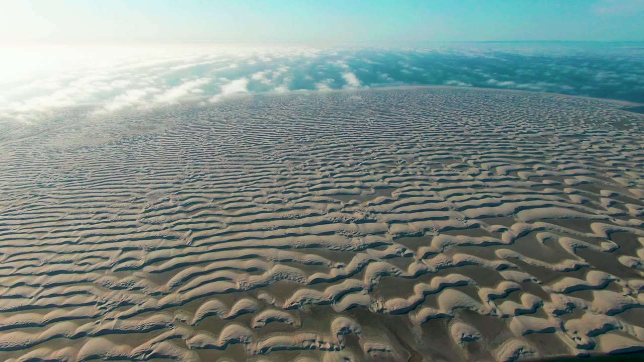 Opening aerial frame over rippled sand dunes in Guerrero Negro, Baja California Sur, with pale cloud bands near the horizon and no visible people.