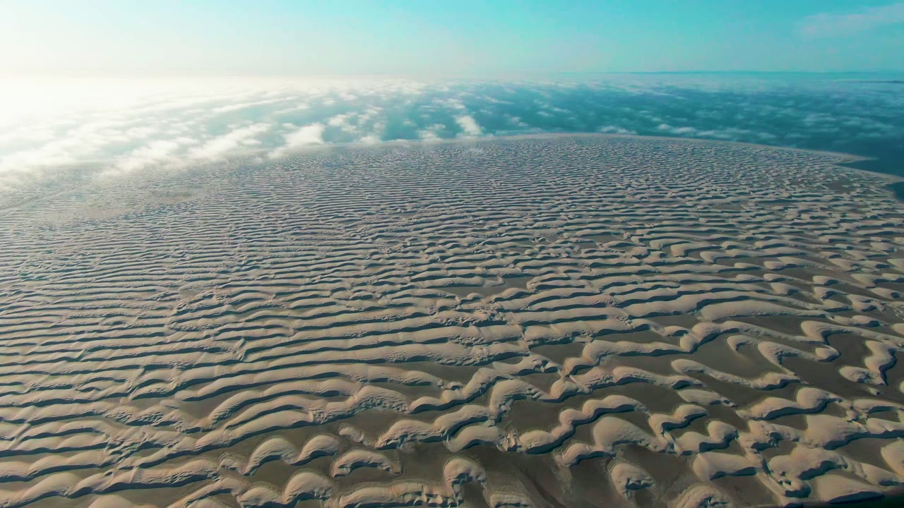Aerial desert sand in Guerrero Negro fills most of the frame, with curved ripple shadows in the foreground, distant cloud streaks, and no visible people.