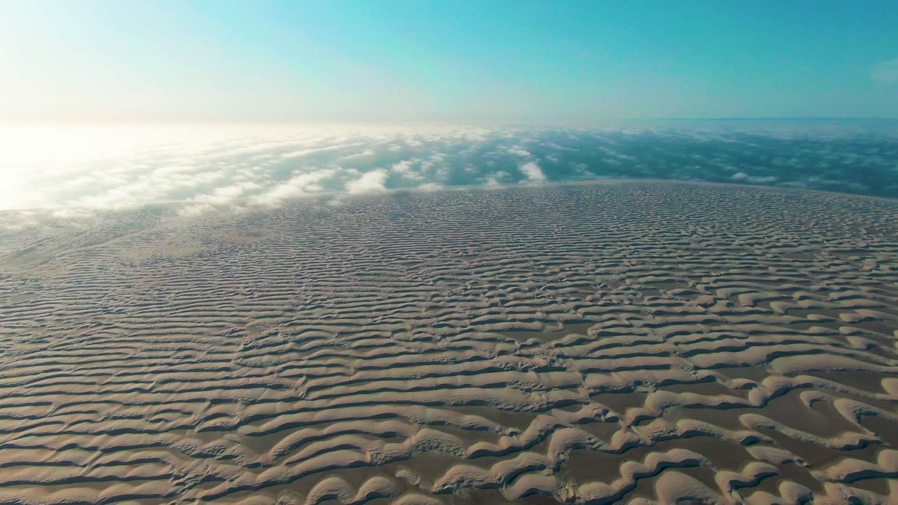 At 0:00, an aerial view over rippled sand dunes in Guerrero Negro, Baja California Sur shows a bright left-side sun glare, a blue sky, distant haze, and no visible people.