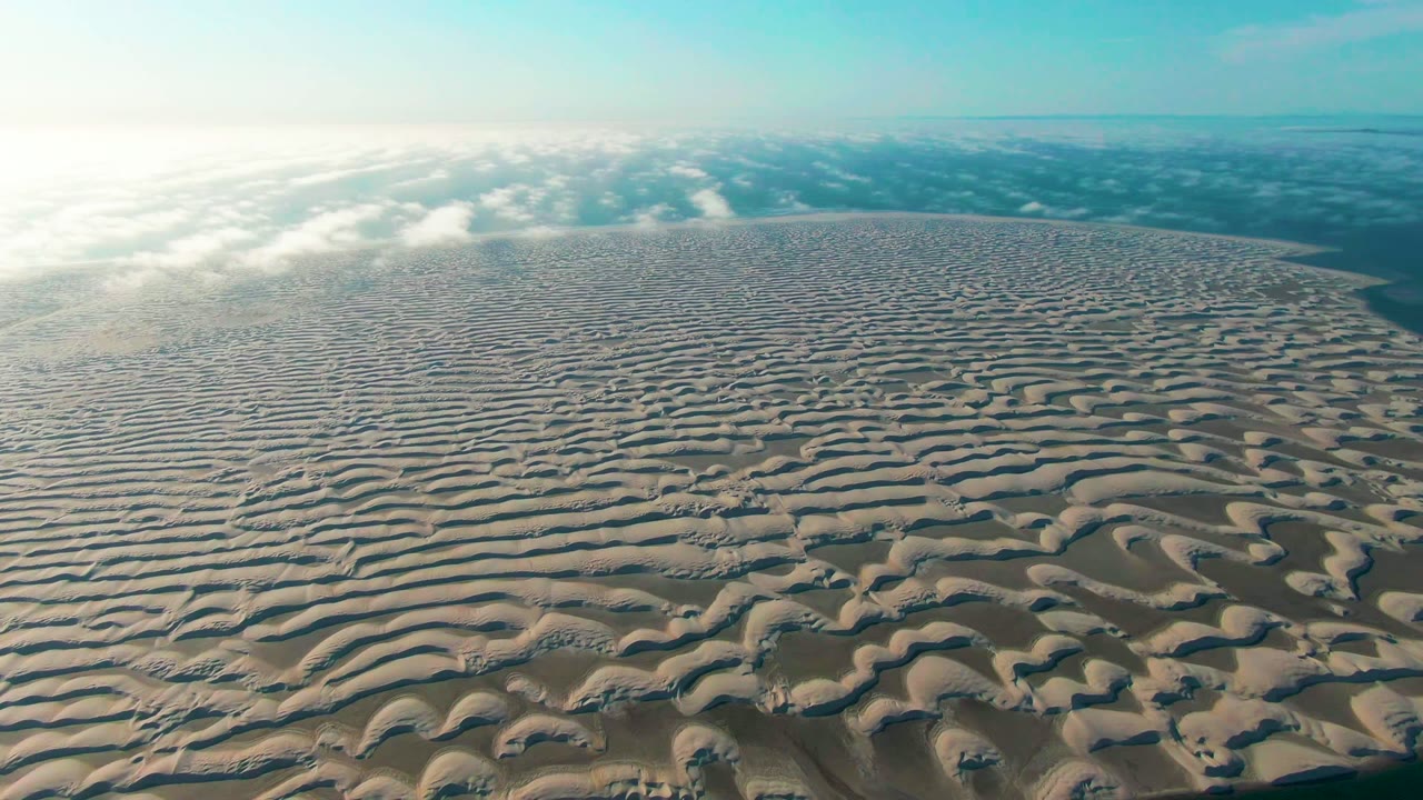 A high aerial view over rippled sand dunes in Guerrero Negro, Baja California Sur, with bright haze at left, cloud bands near the horizon, and no visible people.