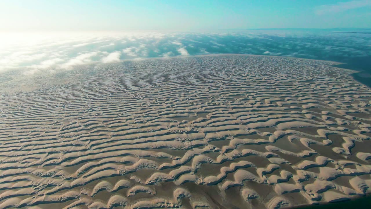 Wide aerial dune field with wavy sand ridges and clouds along the horizon.