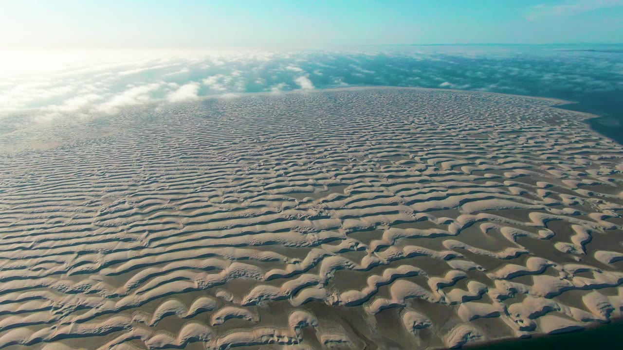 A broad aerial frame of sand ridges in Guerrero Negro shows the dune surface filling most of the image, with soft clouds above and no visible people.