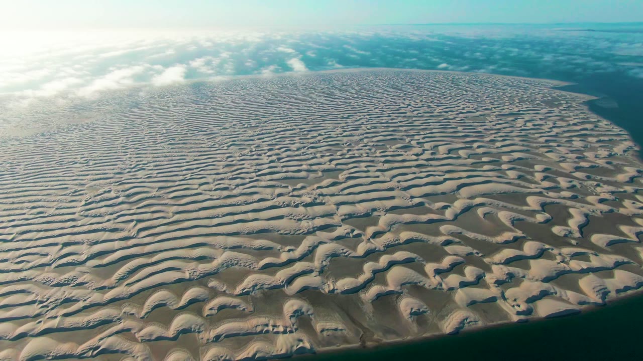 Aerial view over a circular rippled sand dune island in Guerrero Negro, Baja California Sur, with teal water along the lower edge, bright haze at upper left, scattered clouds, and no visible people.