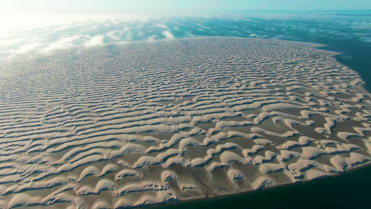 A high oblique aerial frame shows the Guerrero Negro dune island with repeated beige ridges, darker troughs, water curving along the lower right edge, scattered cloud cover, and no visible people.