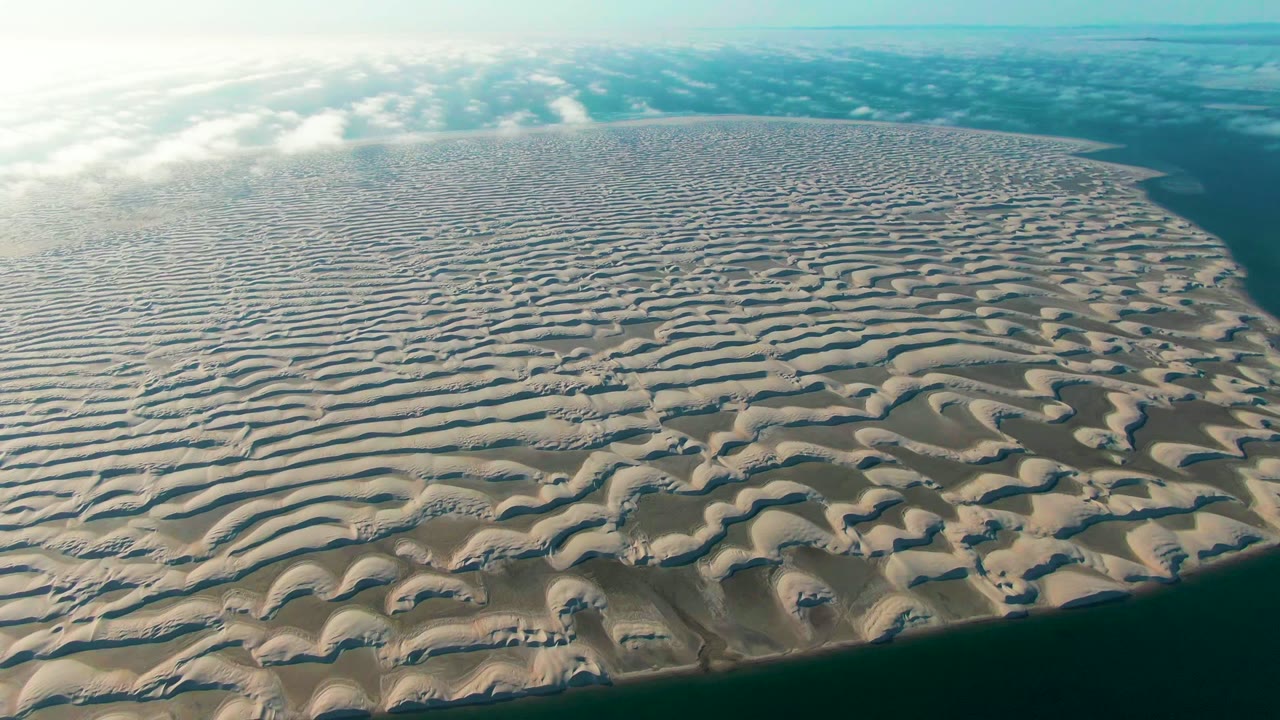 Aerial view of the patterned sand island in Guerrero Negro, Baja California Sur, with dense ripple lines across the center, teal water on the right and bottom edges, cloud shadows near the horizon, and no visible people.