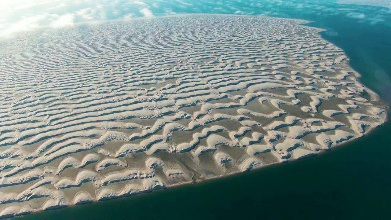 Rippled pale dunes curve toward a dark water channel along the shoreline.