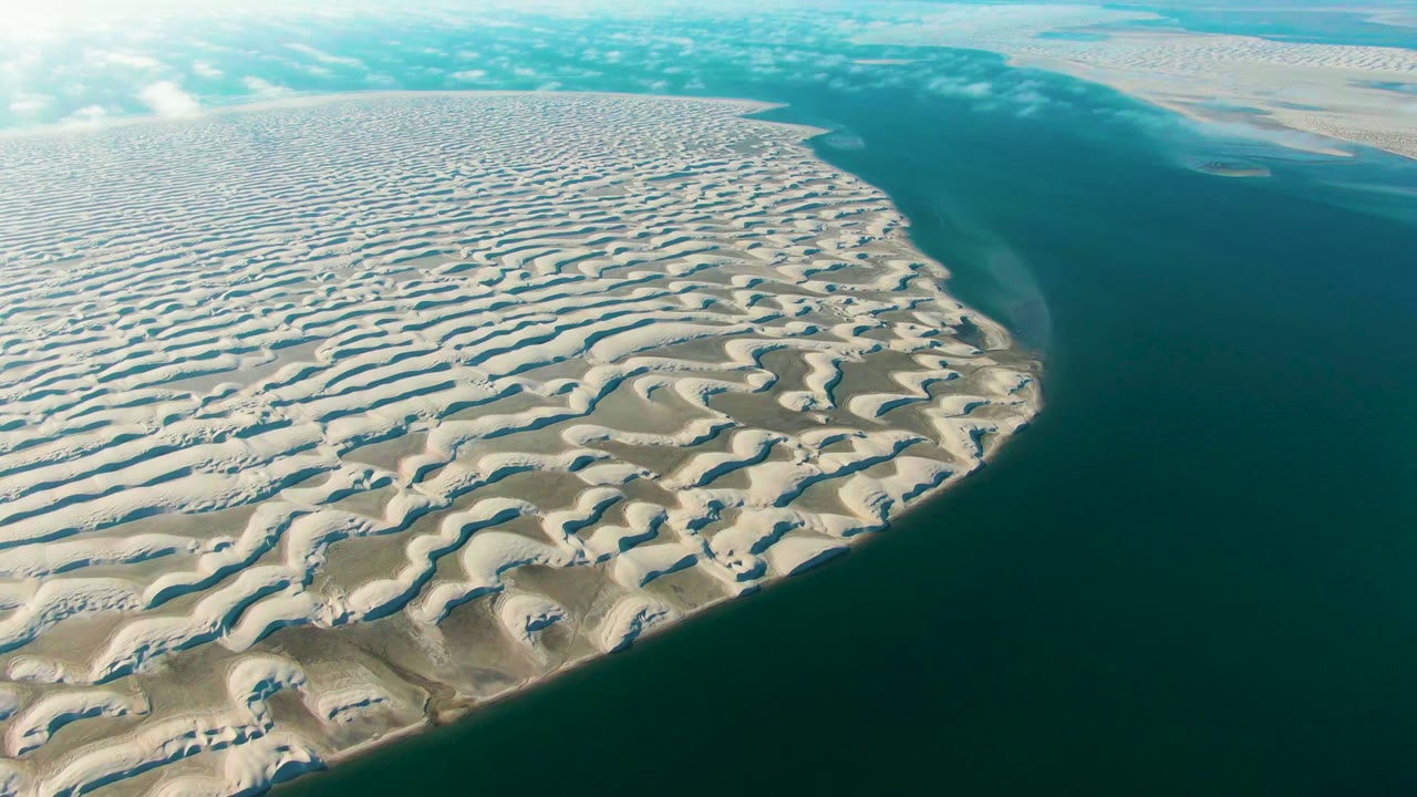 Wide aerial frame of Guerrero Negro coastal dunes, with repeated pale ridges across the left side and teal ocean water on the right, no visible people.