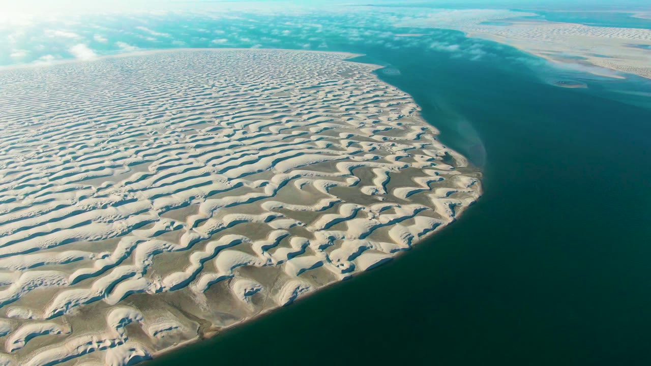 Aerial dune field borders dark calm ocean under scattered clouds.