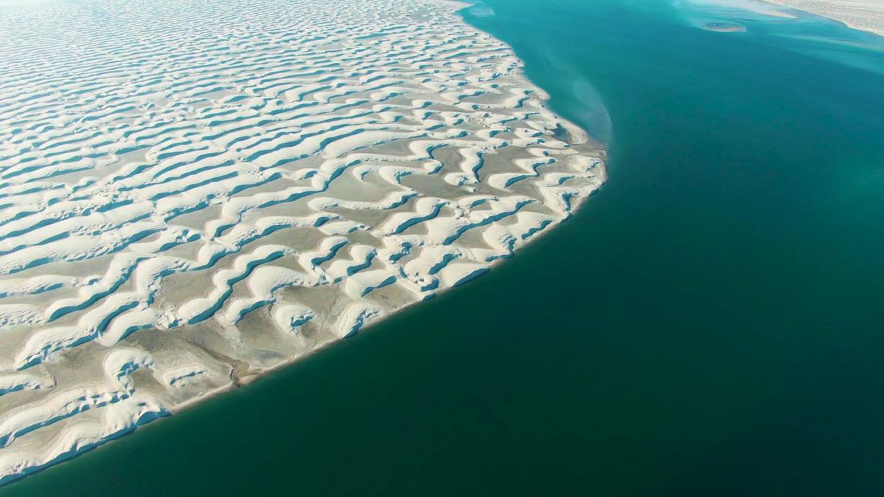 Opening aerial frame over Guerrero Negro, Baja California Sur, with white patterned dunes curving beside turquoise lagoon water and no visible people.