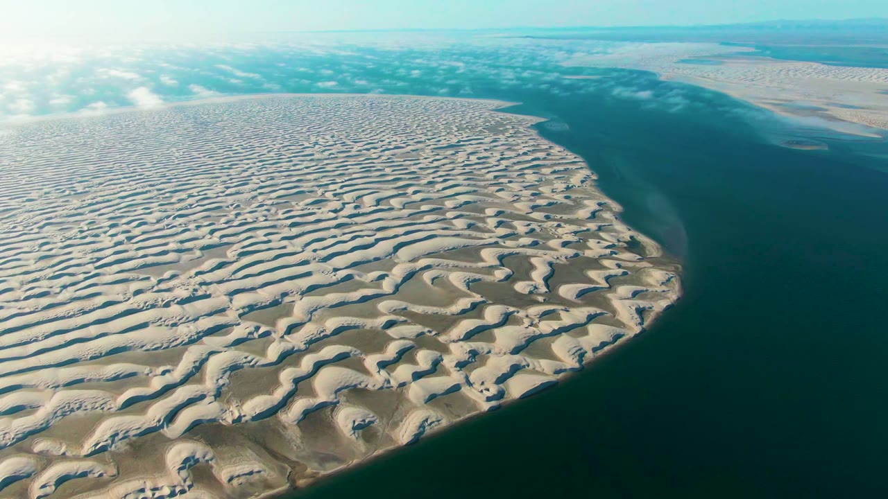 Aerial view over Guerrero Negro sand dunes curving beside dark blue-green ocean water, with pale sky, scattered cloud cover, and no visible people.