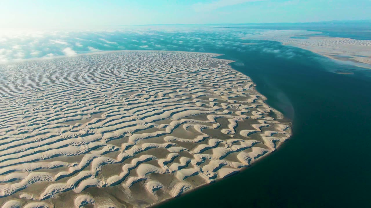 High aerial view in Guerrero Negro with textured sand dunes in the foreground, a dark ocean edge to the right, hazy cloud shadows, and no visible people.