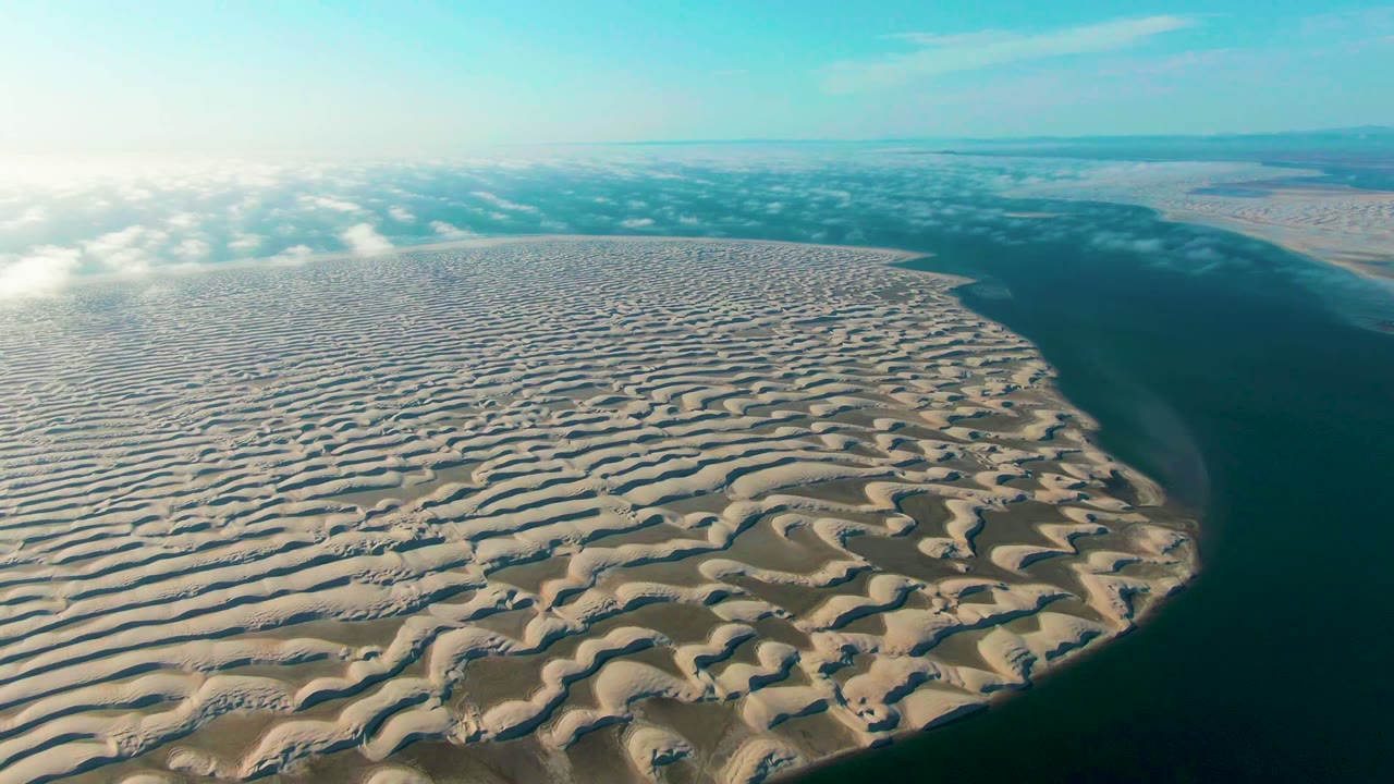 Aerial frame from Guerrero Negro, Baja California Sur, showing patterned beige sand dunes beside dark blue-green ocean water with low clouds on the horizon and no visible people.